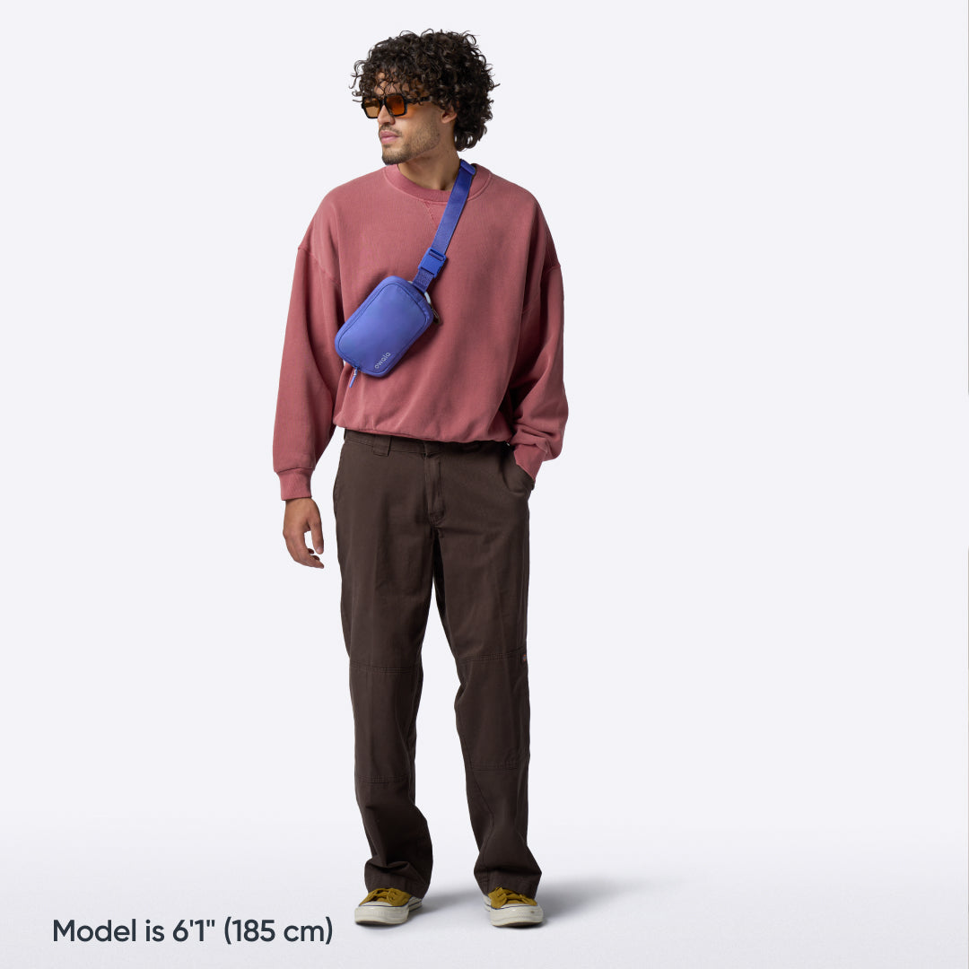 Owala Daily Crossbody in Al Fresco, peach belt bag worn crossbody by a man in a red sweatshirt and brown pants, with text reading "Model is 6'1" (185 cm)"